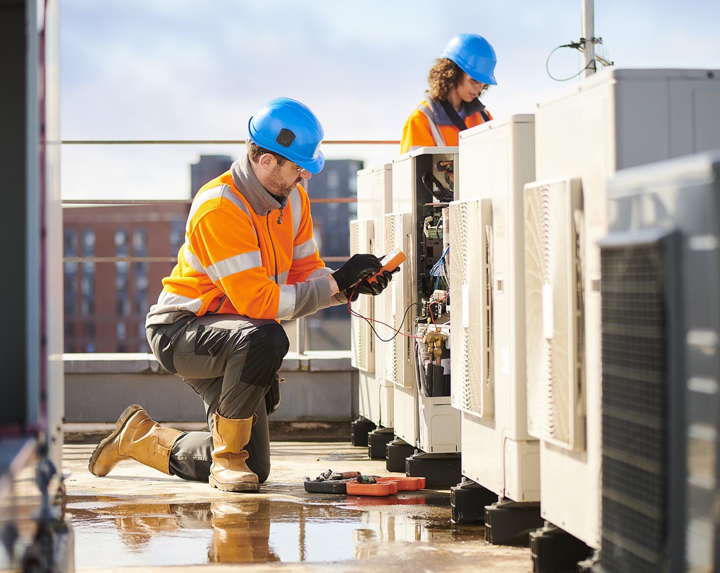 Electricians in hi-vis vests work on HVAC units on top of a building